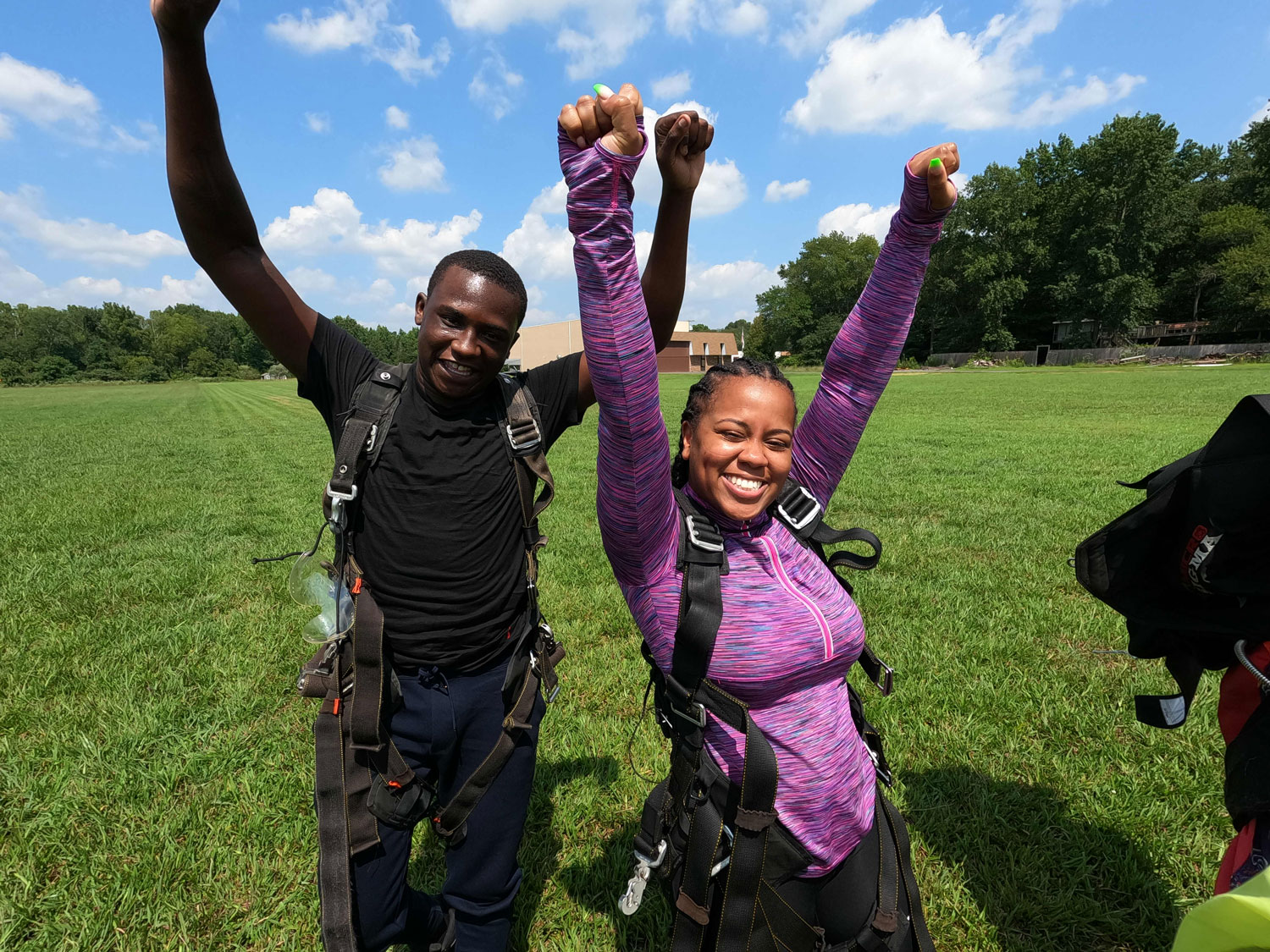 Skydivers smiling and celebrating with arms raised after landing on a sunny field of grass.