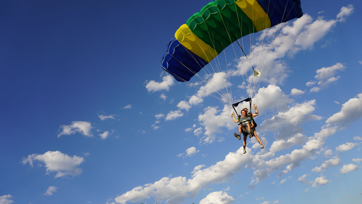 Tandem skydivers in canopy flight under a colorful parachute against a blue sky with clouds.